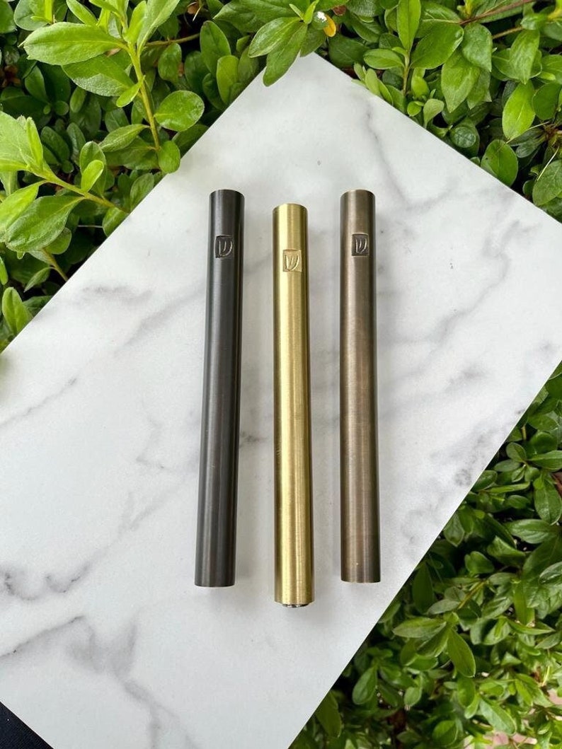 Pode incluir: Tr&ecirc;s canetas met&aacute;licas em diferentes acabamentos: preto, dourado e bronze. Cada caneta tem um logotipo quadrado com a letra "D" no centro.