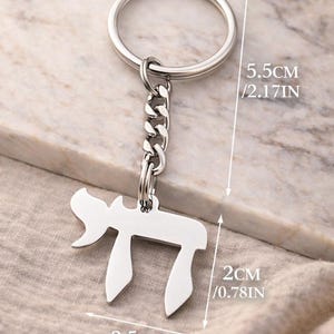 Może przedstawiać: Srebrzysty metalowy brelok do kluczy z hebrajską literą Chai. Całkowita długość wynosi 5,5 cm. Zawieszka ma 2 cm wysokości i 2,5 cm szerokości i jest przymocowana do standardowego okrągłego k&oacute;łka. Wymiary są wyraźnie zaznaczone na zdjęciu.