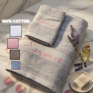 May include: A set of light gray 100% cotton towels featuring the name Emma embroidered in pink script. The set includes a smaller hand towel and a larger bath towel, accompanied by a small glass bottle of oil, a bar of soap, and lavender sprigs.