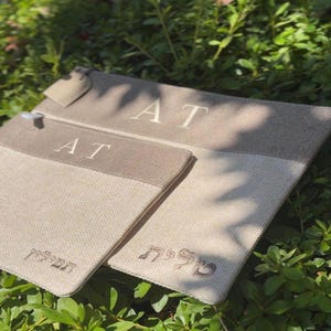 May include: Two rectangular beige fabric pouches with a darker beige horizontal band at the top. The pouches feature the letters AT embroidered in white on the band. The smaller pouch has Hebrew text embroidered in white, and the larger has Hebrew text.