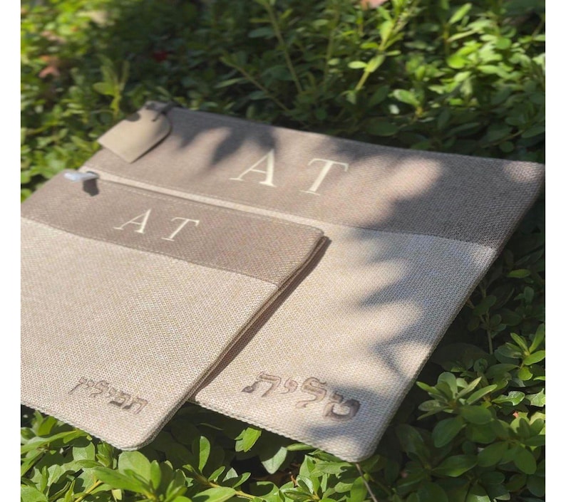 May include: Two rectangular beige fabric pouches with a darker beige horizontal band at the top. The pouches feature the letters AT embroidered in white on the band. The smaller pouch has Hebrew text embroidered in white, and the larger has Hebrew text.