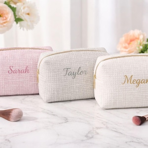 Op de afbeelding: Drie rechthoekige make-uptasjes met een textuurstof afwerking. De roze tas heeft de naam Sarah, de grijze tas heeft de naam Taylor en de crèmekleurige tas heeft de naam Megan, allemaal geborduurd in cursief schrift met gouden ritsen.