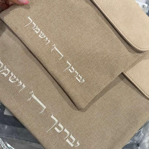 May include: A beige textured fabric bag featuring a smooth flap and white embroidered Hebrew text that reads "יְבָרֶכְךָ ה' וְיִשְׁמְרֶךָ". The bag has a structured rectangular shape with a clean finish and a matching beige flap closure at the top.