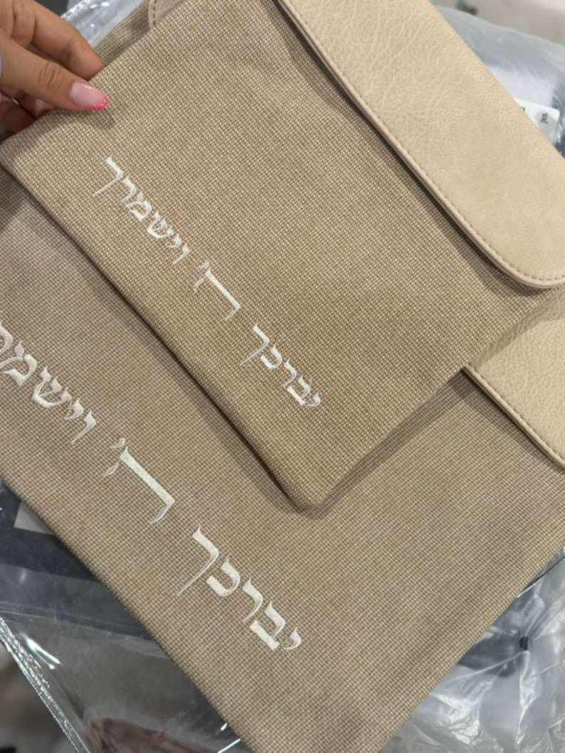 May include: A beige textured fabric bag featuring a smooth flap and white embroidered Hebrew text that reads "יְבָרֶכְךָ ה' וְיִשְׁמְרֶךָ". The bag has a structured rectangular shape with a clean finish and a matching beige flap closure at the top.