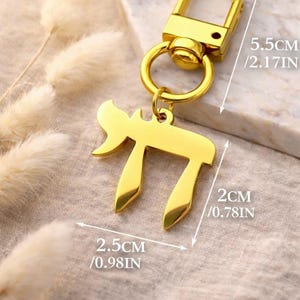 Może przedstawiać: Brelok w kolorze złota z hebrajskim symbolem Chai. Całkowita długość wynosi 5,5 cm, a zawieszka ma 2,5 cm szerokości i 2 cm wysokości. Brelok posiada obrotowe zapięcie typu karabińczyk oraz okrągłe k&oacute;łko na klucze.
