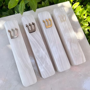 May include: Four white mezuzah cases with a marbled appearance. Each case features a different colored Hebrew letter Shin. The Shin letters are in silver, dark brown, and gold. The cases are rectangular with rounded edges.