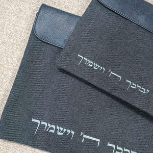 May include: Two dark gray rectangular fabric cases with dark blue faux leather flaps. Each case features white embroidered Hebrew text that reads יְבָרֶכְךָ ה' וְיִשְׁמְרֶךָ. The fabric has a fine, textured weave pattern, and the flaps have a smooth, grained finish.
