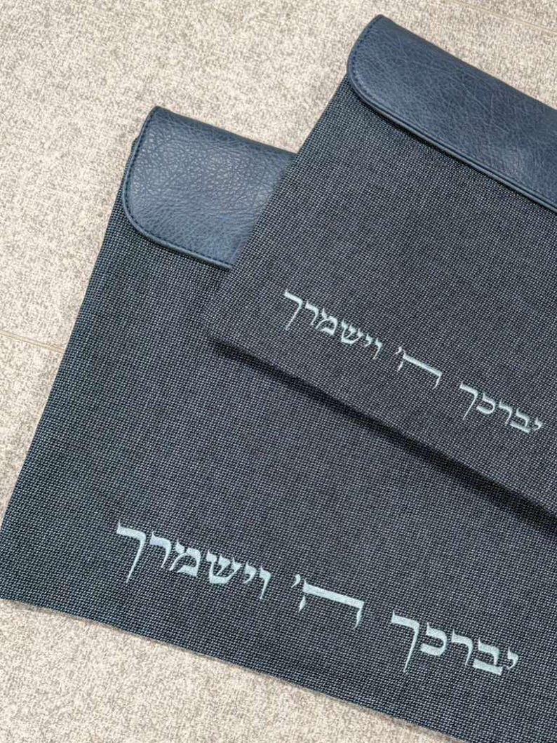 May include: Two dark gray rectangular fabric cases with dark blue faux leather flaps. Each case features white embroidered Hebrew text that reads יְבָרֶכְךָ ה' וְיִשְׁמְרֶךָ. The fabric has a fine, textured weave pattern, and the flaps have a smooth, grained finish.