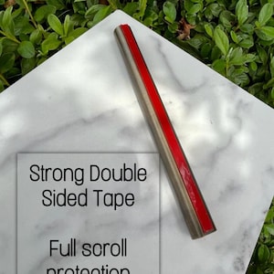 Pode incluir: Um rolo de fita adesiva de dupla face resistente sobre uma superf&iacute;cie de m&aacute;rmore branco. A fita &eacute; vermelha e tem um suporte prateado. O texto "Strong Double Sided Tape" e "Full scroll protection" &eacute; vis&iacute;vel na imagem.