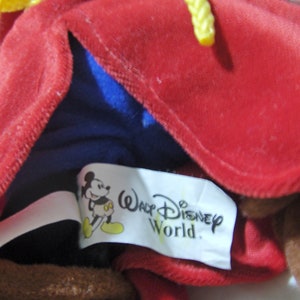 May include: A red plush toy with a yellow rope tied around the neck. The toy is wearing a blue shirt and has a white tag that says "Walt Disney World" with a cartoon Mickey Mouse image.