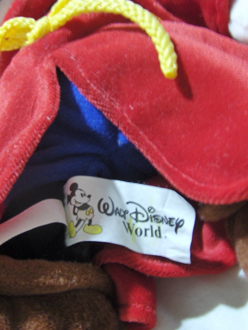 May include: A red plush toy with a yellow rope tied around the neck. The toy is wearing a blue shirt and has a white tag that says "Walt Disney World" with a cartoon Mickey Mouse image.