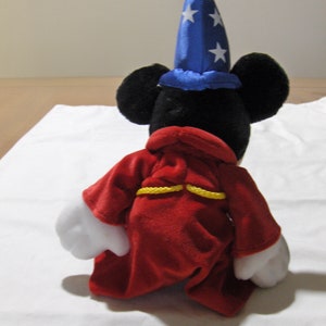 May include: A plush Mickey Mouse toy wearing a blue and white star-patterned hat and a red robe with yellow trim. The toy is sitting on a white surface.