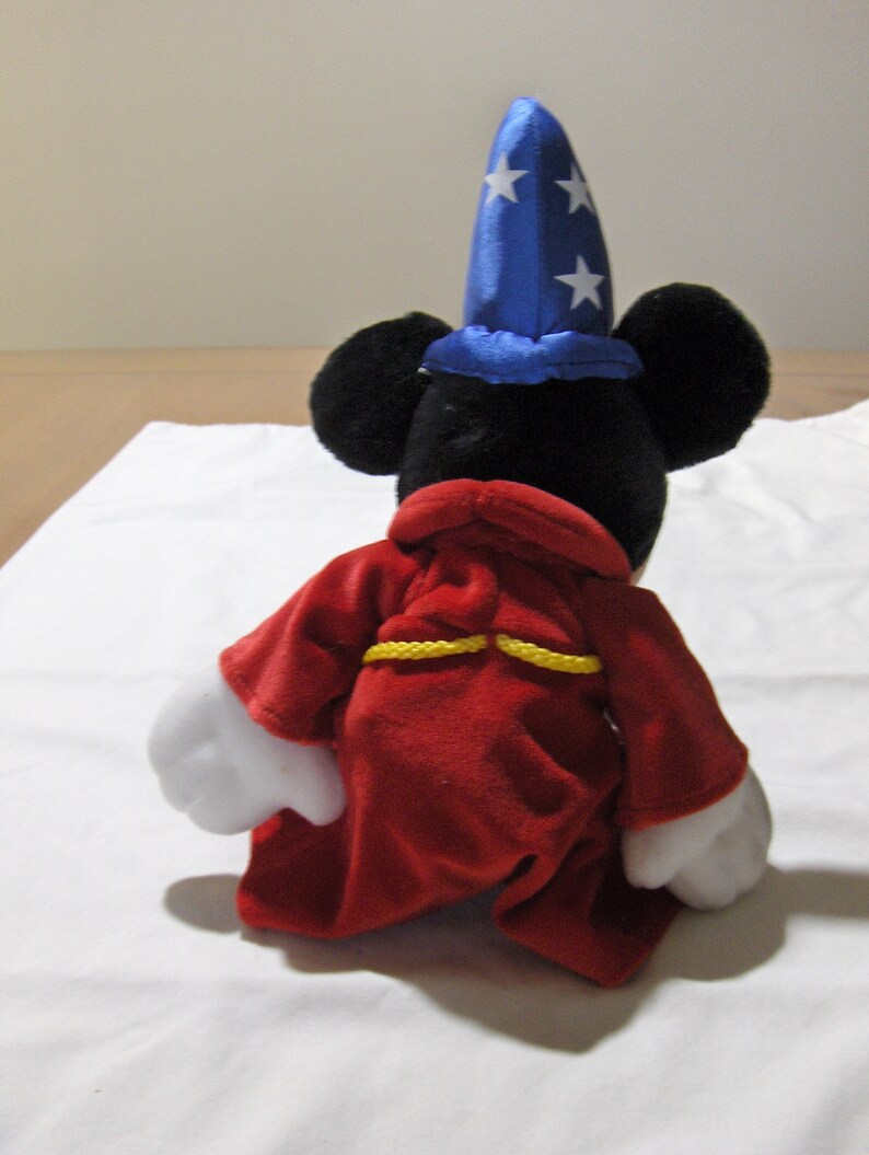 May include: A plush Mickey Mouse toy wearing a blue and white star-patterned hat and a red robe with yellow trim. The toy is sitting on a white surface.