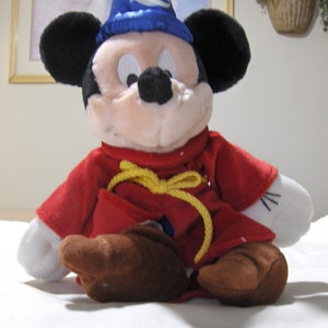 May include: A plush Mickey Mouse toy wearing a blue wizard hat with a yellow star and a red robe with a yellow rope belt.