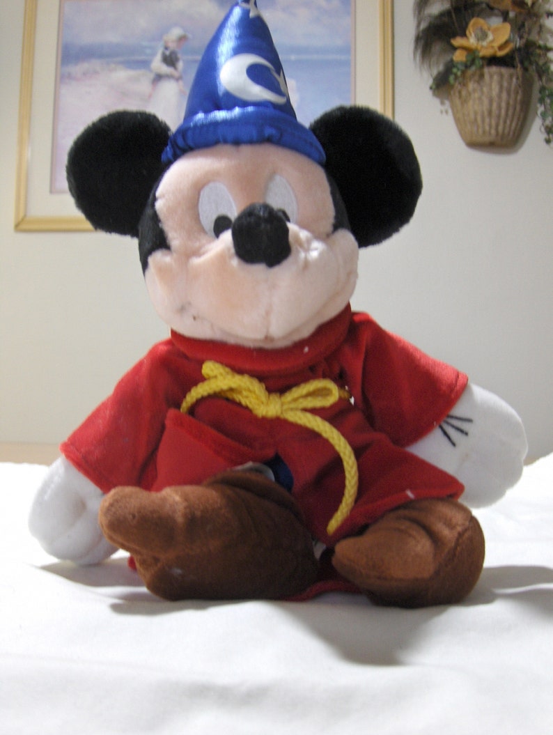 May include: A plush Mickey Mouse toy wearing a blue wizard hat with a yellow star and a red robe with a yellow rope belt.
