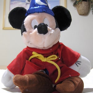 May include: A plush Mickey Mouse toy wearing a blue wizard hat with a crescent moon design and a red robe with a yellow rope belt.