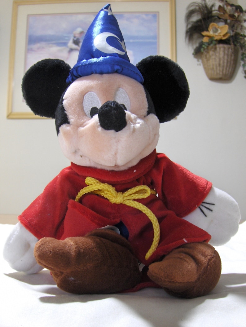 May include: A plush Mickey Mouse toy wearing a blue wizard hat with a crescent moon design and a red robe with a yellow rope belt.