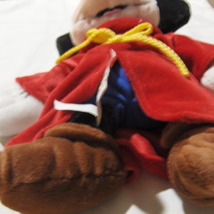 May include: A plush Mickey Mouse toy wearing a red robe with a yellow sash and blue lining. The toy has brown fur and black ears.