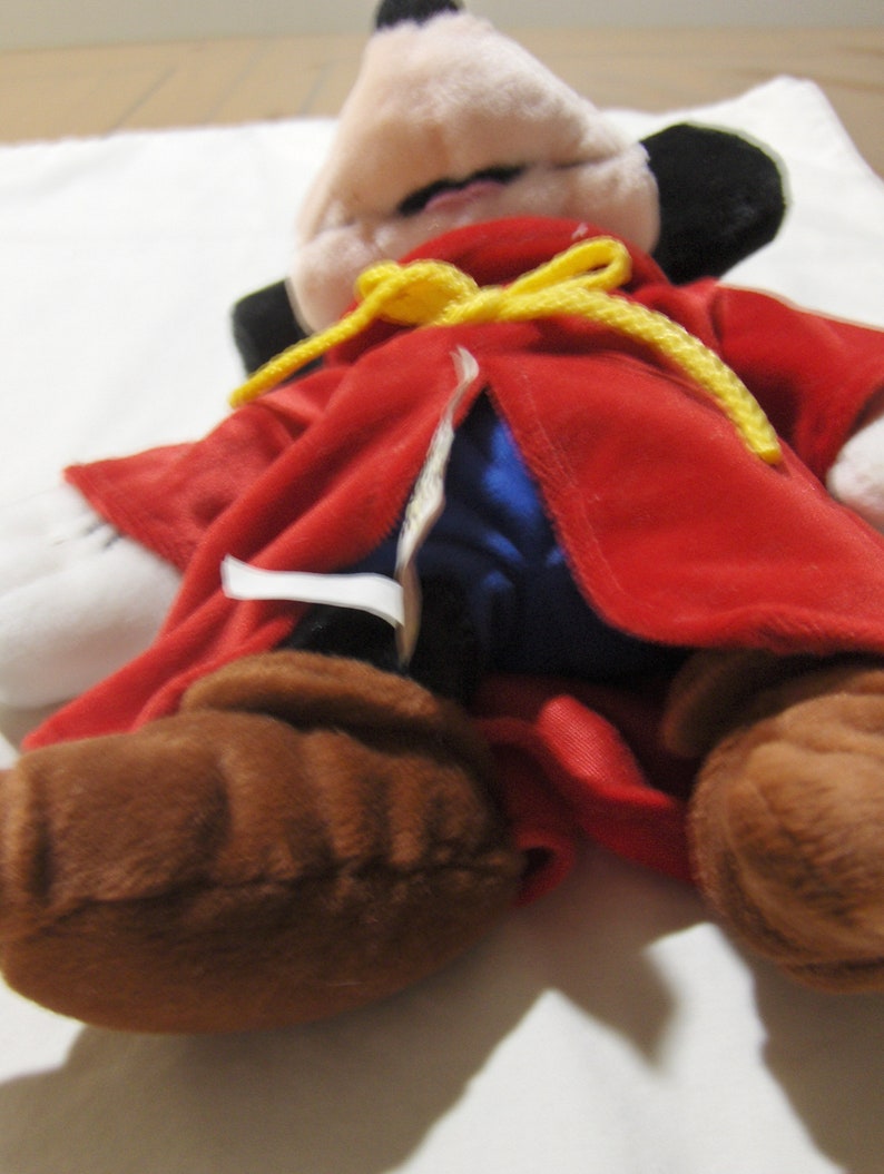 May include: A plush Mickey Mouse toy wearing a red robe with a yellow sash and blue lining. The toy has brown fur and black ears.