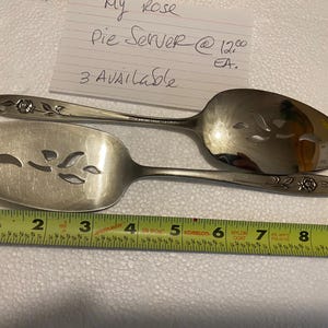 May include: Two silver pie servers with floral detailing. One server has a slotted blade with leaf-shaped cutouts, and the other is a serving spoon. A tape measure shows the length of the servers. The text "My Rose Pie Server" is visible.
