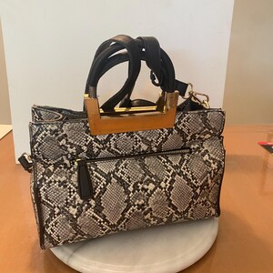 May include: A snakeskin-patterned handbag with black handles and gold-colored hardware. The bag has a rectangular shape with a zipper closure and a detachable shoulder strap. The bag is sitting on a white, round surface.