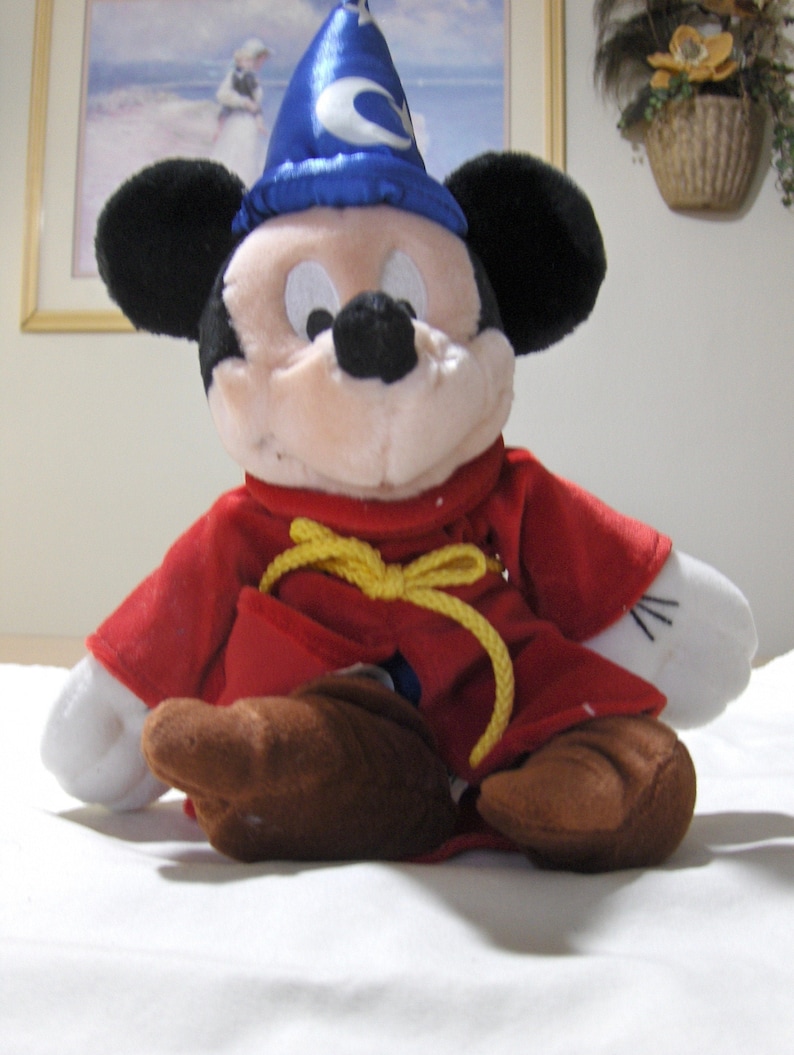 May include: A plush Mickey Mouse toy wearing a blue wizard hat with a yellow rope tied around his neck and a red robe.