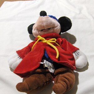 May include: A red plush Mickey Mouse dressed as a sorcerer with a yellow sash and brown boots. The plush toy has a blue hat and white gloves. The tag on the toy reads "Disney World".