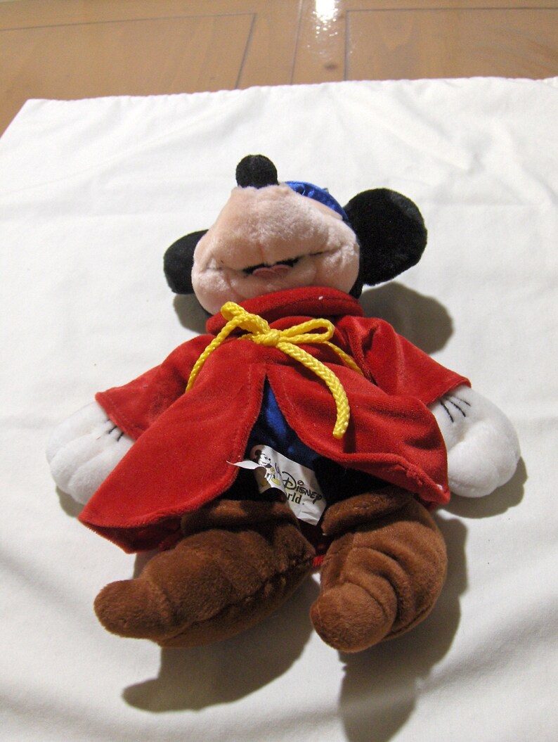 May include: A red plush Mickey Mouse dressed as a sorcerer with a yellow sash and brown boots. The plush toy has a blue hat and white gloves. The tag on the toy reads "Disney World".