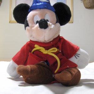 May include: A plush Mickey Mouse toy wearing a blue wizard hat with a yellow crescent moon. The toy is wearing a red robe with a yellow sash and has brown shoes.