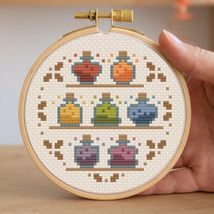 Könnte beinhalten: Ein Kreuzstich-Stickrahmen mit einem Design aus bunten Zaubertränken. Die Flaschen sind auf Regalen angeordnet und weisen verschiedene Farben auf, darunter Rot, Orange, Gelb, Grün, Blau, Lila und Rosa. Die Stickerei ist in einem Holzrahmen gefasst.