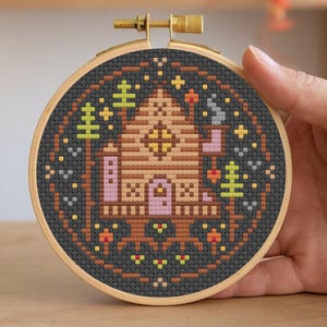 May include: A cross-stitch embroidery hoop with a dark background featuring a brown cabin with a pink door and yellow windows. The cabin is surrounded by trees and stars.