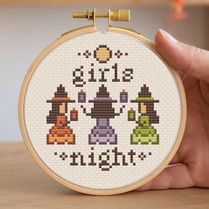 May include: A cross-stitch embroidery hoop featuring the words "girls night" and three pixelated witch figures. The witches are in orange, purple, and green dresses, holding candles. The hoop is held in a hand.
