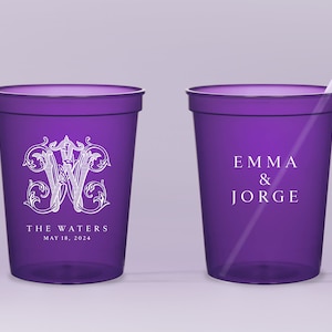 May include: Two purple plastic cups with white text. One cup says "The Waters May 18, 2024" with a decorative monogram. The other cup says "Emma & Jorge".