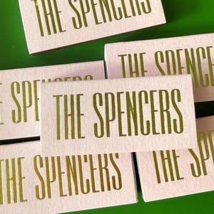 May include: A collection of rectangular light pink cards featuring the text THE SPENCERS printed in a metallic gold font. Each card displays the same name in a tall, sans-serif typeface, arranged in a scattered overlapping pattern on a green surface.
