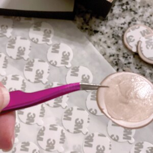 May include: A pair of tweezers holding a round, light brown adhesive sticker with a shiny, slightly textured surface. The sticker is surrounded by many white, round, adhesive stickers with the text "3M" printed on them.