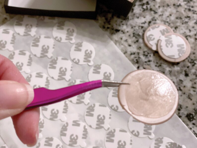 May include: A pair of tweezers holding a round, light brown adhesive sticker with a shiny, slightly textured surface. The sticker is surrounded by many white, round, adhesive stickers with the text "3M" printed on them.