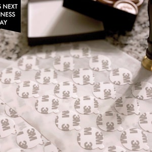 May include: A close-up of a sheet of white paper with multiple round, white adhesive stickers. The stickers have the 3M logo printed on them. The text "SHIPS NEXT BUSINESS DAY" is visible in the top left corner of the image.