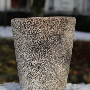 Könnte beinhalten: Eine strukturierte, zylindrische Vase mit einer gesprenkelten, porösen Oberfläche. Die Vase ist überwiegend grau mit weißen und silbernen Akzenten und steht auf einer schneebedeckten Oberfläche. Das Design verleiht der Vase ein einzigartiges, organisches Aussehen, geeignet für die Inneneinrichtung.