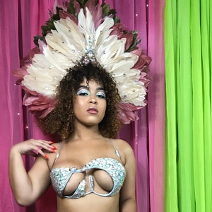 May include: A woman wearing a light blue, sequined, two-piece costume with a large feathered headdress. The headdress is pink and white with rhinestones. The woman is standing in front of a pink and green curtain.