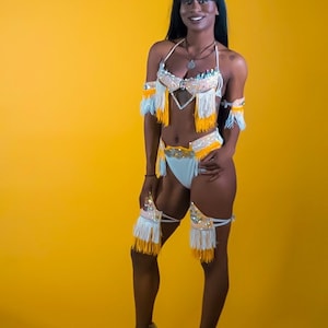 May include: A woman wearing a white and yellow costume with fringe and feathers. She is wearing a white bra with yellow fringe and a white skirt with yellow fringe. She is also wearing a white headpiece with yellow feathers. She is standing on a yellow background.