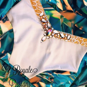 May include: A white satin bodice with a gold and rhinestone embellishment. The embellishment features a large, teardrop-shaped rhinestone in the center, surrounded by smaller rhinestones and gold sequins. The bodice is labeled "B. Royale Designs".
