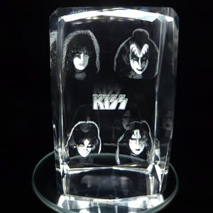 May include: A clear, rectangular crystal block featuring etched images of the rock band KISS. The band's logo is visible, along with portraits of the band members. The block sits on a clear, circular base.