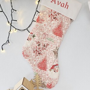May include: A Christmas stocking with a white cuff embroidered with the name "Avah" in red. The stocking features a festive pattern of red trees, Santa Claus, and reindeer on a cream background. A wooden Christmas house is in the foreground.