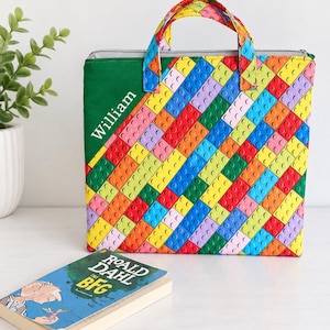 May include: A rectangular fabric tote bag featuring a colourful diagonal pattern of interlocking plastic building blocks in red, blue, yellow, green, and purple. The bag has a solid green corner panel with the name William printed in white text.