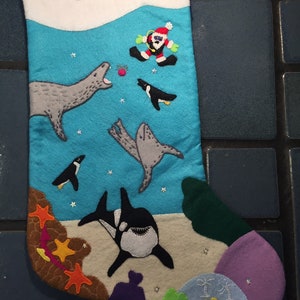 May include: A blue and white Christmas stocking with the words "Merry Christmas" embroidered on the top. The stocking features a variety of sea creatures, including a penguin, a seal, an orca, and a Santa Claus figure.