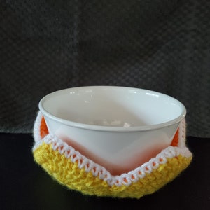 May include: A white ceramic bowl is sitting inside a crocheted bowl cozy. The cozy is yellow with a white and orange trim.