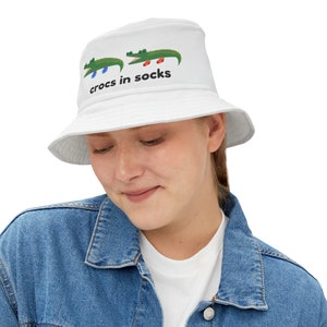 Puede incluir: Sombrero de pescador blanco con dos caimanes verdes de dibujos animados, pies azules y rojos, y el texto "crocs in socks" en negro. El sombrero está hecho de un material suave.