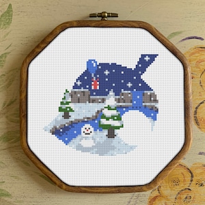 May include: A framed cross-stitch design depicting a winter scene with a house, trees, and a snowman. The house is blue and white with a red roof and a chimney. The trees are green and white. The snowman is white with a black hat and a carrot nose.