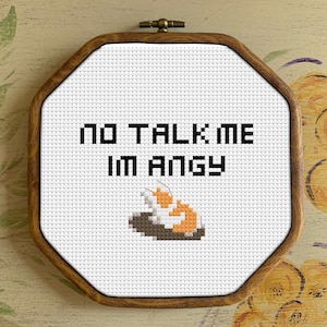 May include: A white cross-stitch embroidery hoop with a brown wooden frame. The embroidery features a cat with orange and white fur, curled up on a black surface. The text "NO TALK ME IM ANGY" is stitched in black thread above the cat.