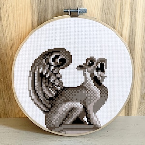 May include: A cross-stitch embroidery hoop with a gray and white image of a gargoyle with a curled tail and wings.
